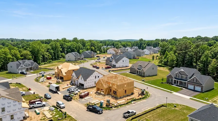 An aerial view of a new suburban residential development with modern houses at various construction stages, surrounded by green trees.