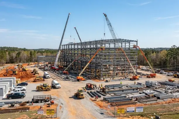 A sprawling construction site with a massive multi-story steel frame, multiple cranes, and heavy machinery under a clear sky.
