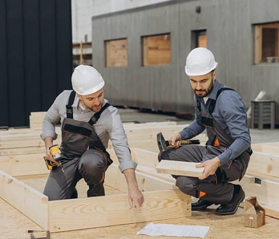 Two carpenters working on a wooden frame in a workshop, using hammer and screwdriver, for prefabricated modular building construction